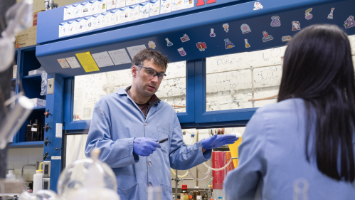 A professor and student wearing blue coats talk in a pharmacy lab