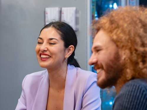 Two coworkers in an office room smile at something out of frame