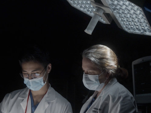 A faculty member and resident in a dark operating room look down at something out of frame