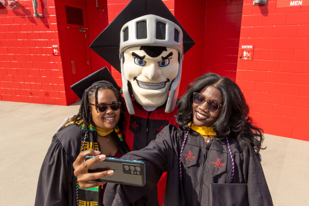 Two graduates taking a selfie pic with the Scarlet Knight mascot