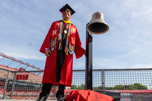 A graduate posing next to the Red Lion Bell at commencement